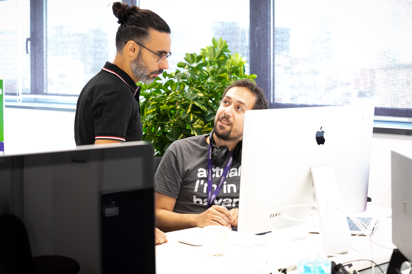 Two male employees talk at a desk in front of a screen